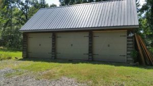 A three-car garage with a metal roof stands on a grassy lot, surrounded by trees. The garage doors are closed, and the structure has a rustic wooden exterior. Sunlight filters through the trees in the background.