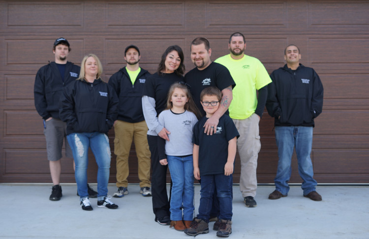 DSC06120 A group of eight adults and two children stand smiling in front of a large brown garage door. Most adults wear black jackets; one wears a bright yellow shirt. The two children stand at the front, side by side.