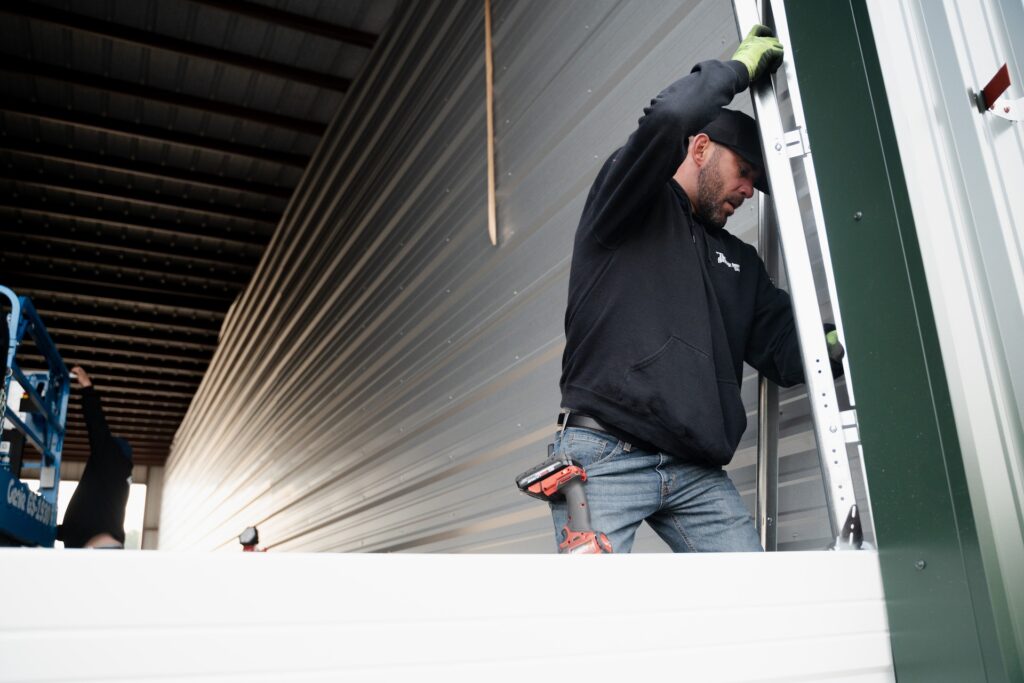 A man wearing a black hoodie and gloves climbs a metal ladder inside a large industrial building, ready for winter maintenance with a power drill strapped to his jeans. The space features metal siding and a high, open ceiling.